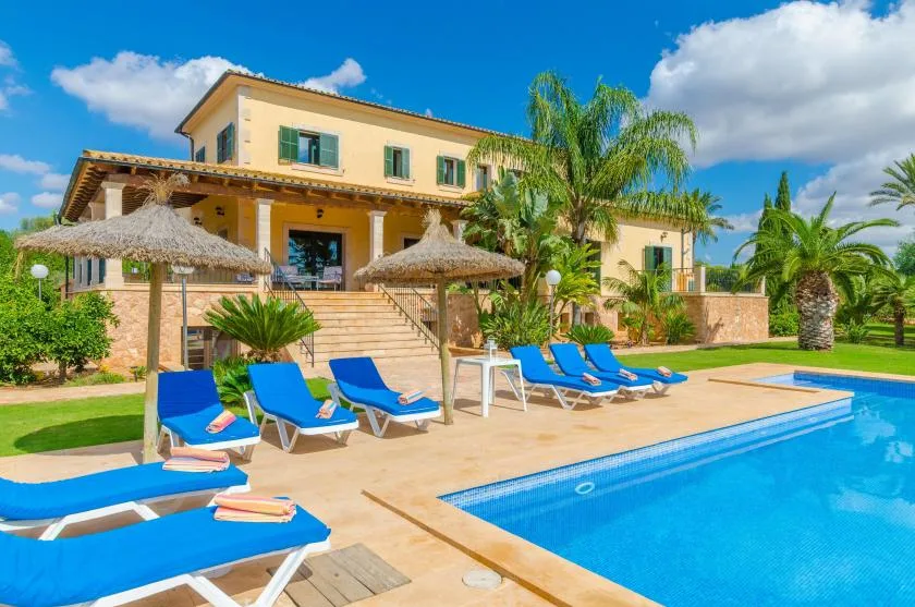 Una casa grande da a una piscina con tumbonas y sombrillas de paja.