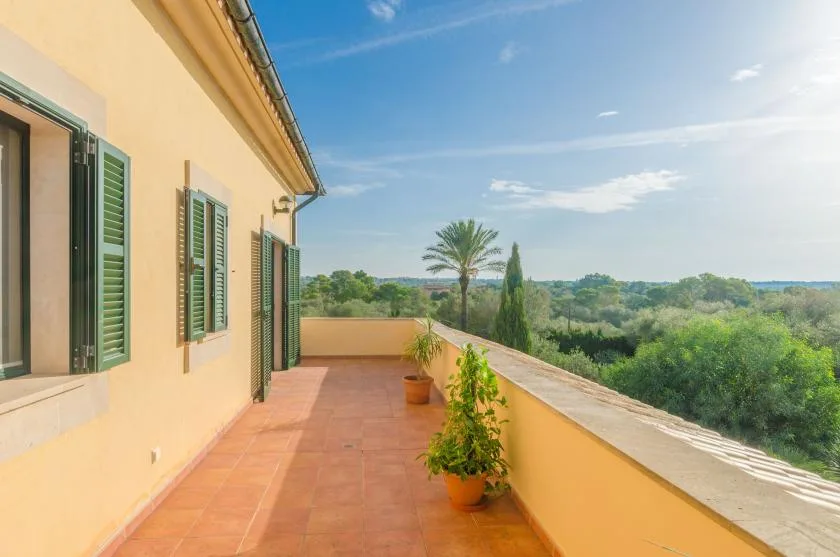 Una terraza soleada junto a una casa mira hacia árboles verdes y colinas lejanas.