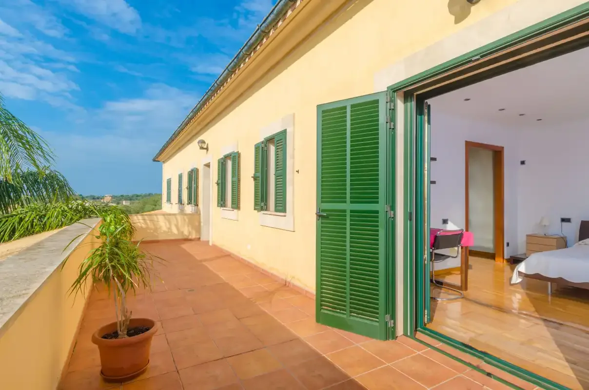 Una terraza soleada con contraventanas verdes se abre a un dormitorio luminoso.
