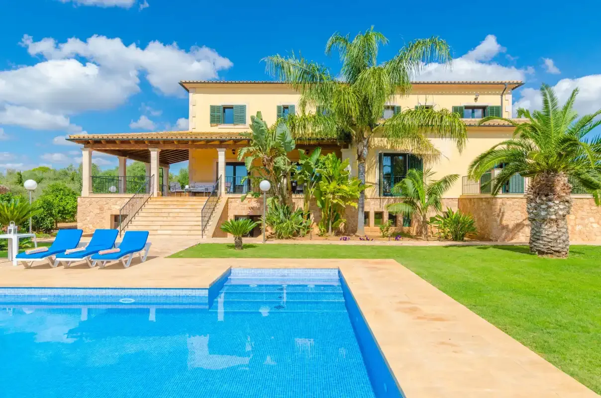 Una villa se alza junto a una piscina, con palmeras y tumbonas cerca.