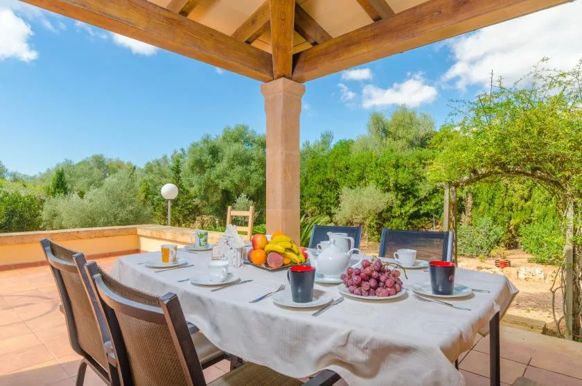 Una mesa de comedor en el patio está puesta con fruta y platos bajo una pérgola de madera.