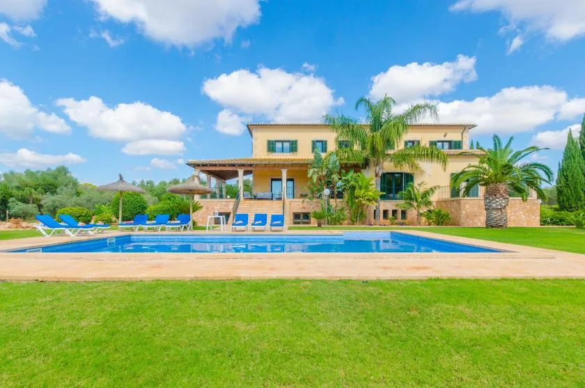 Una piscina y un césped están frente a una casa de dos plantas bajo un cielo azul.