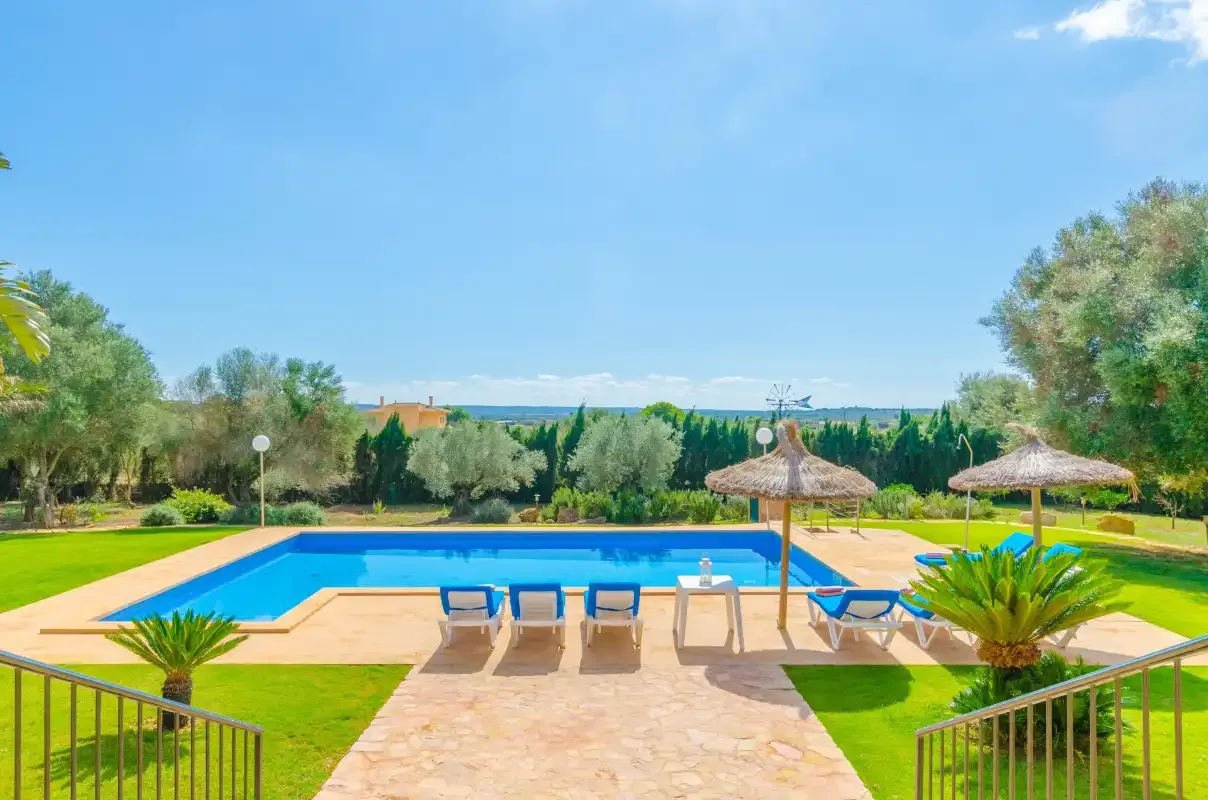 Una piscina al aire libre está rodeada por un patio soleado y césped verde.