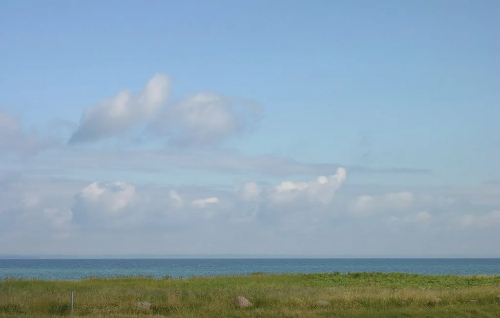 A coastal landscape with grassy dunes, blue sea, and a cloudy sky.