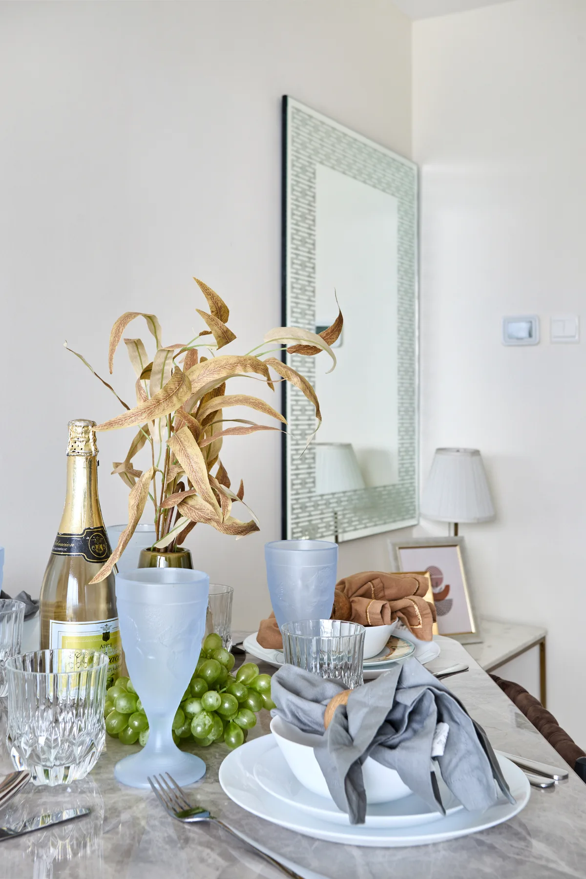 Una mesa de comedor está preparada con platos, servilletas dobladas, cristalería y una planta decorativa.