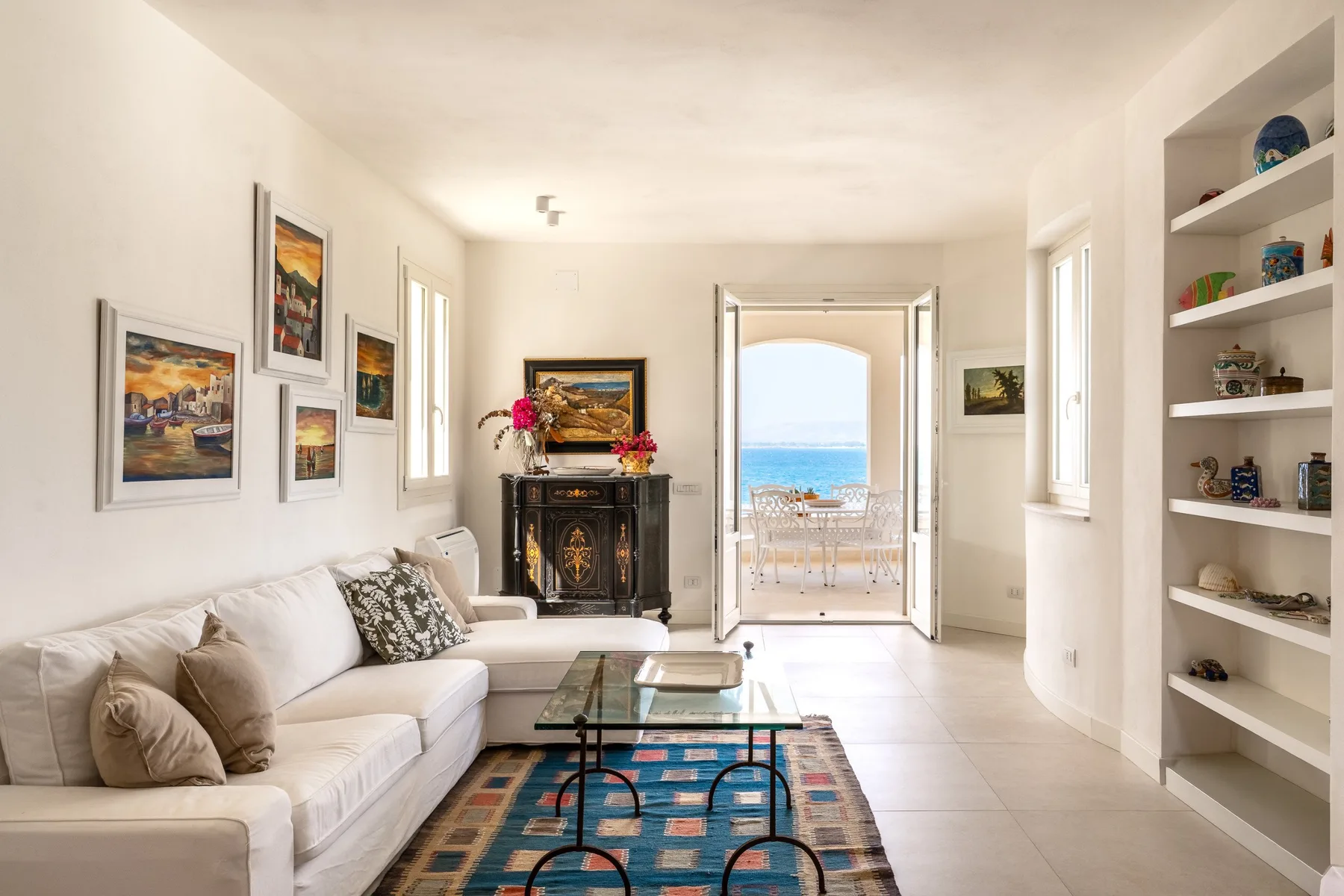 Una sala luminosa con asientos, una mesa de centro de vidrio y puertas abiertas hacia el mar.