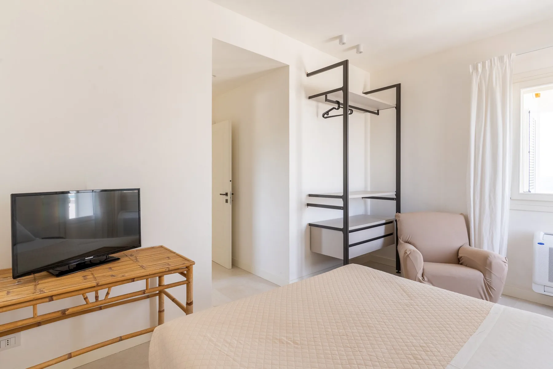 Un dormitorio luminoso con televisor, perchero abierto y asiento tapizado.
