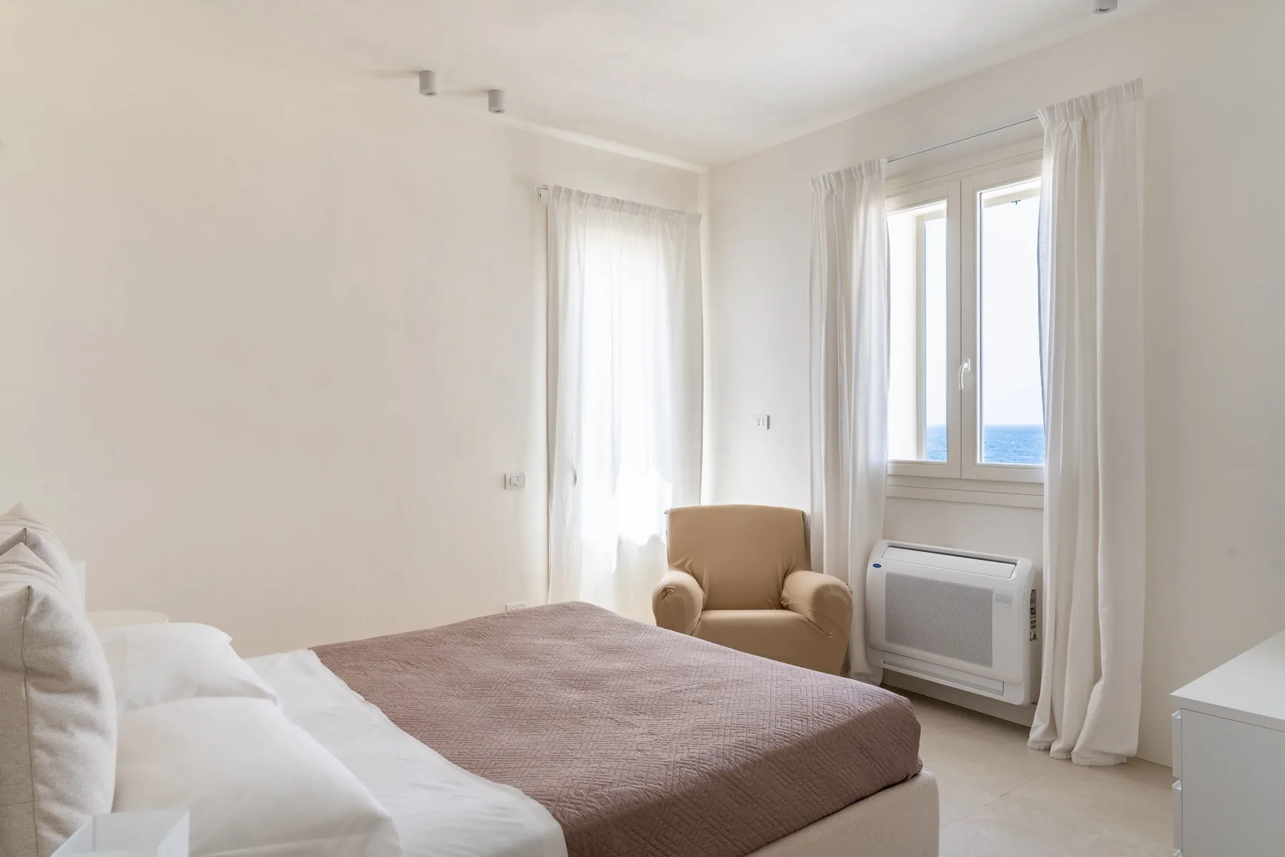 Un dormitorio luminoso con una cama, un sillón y ventanas con vistas al agua.