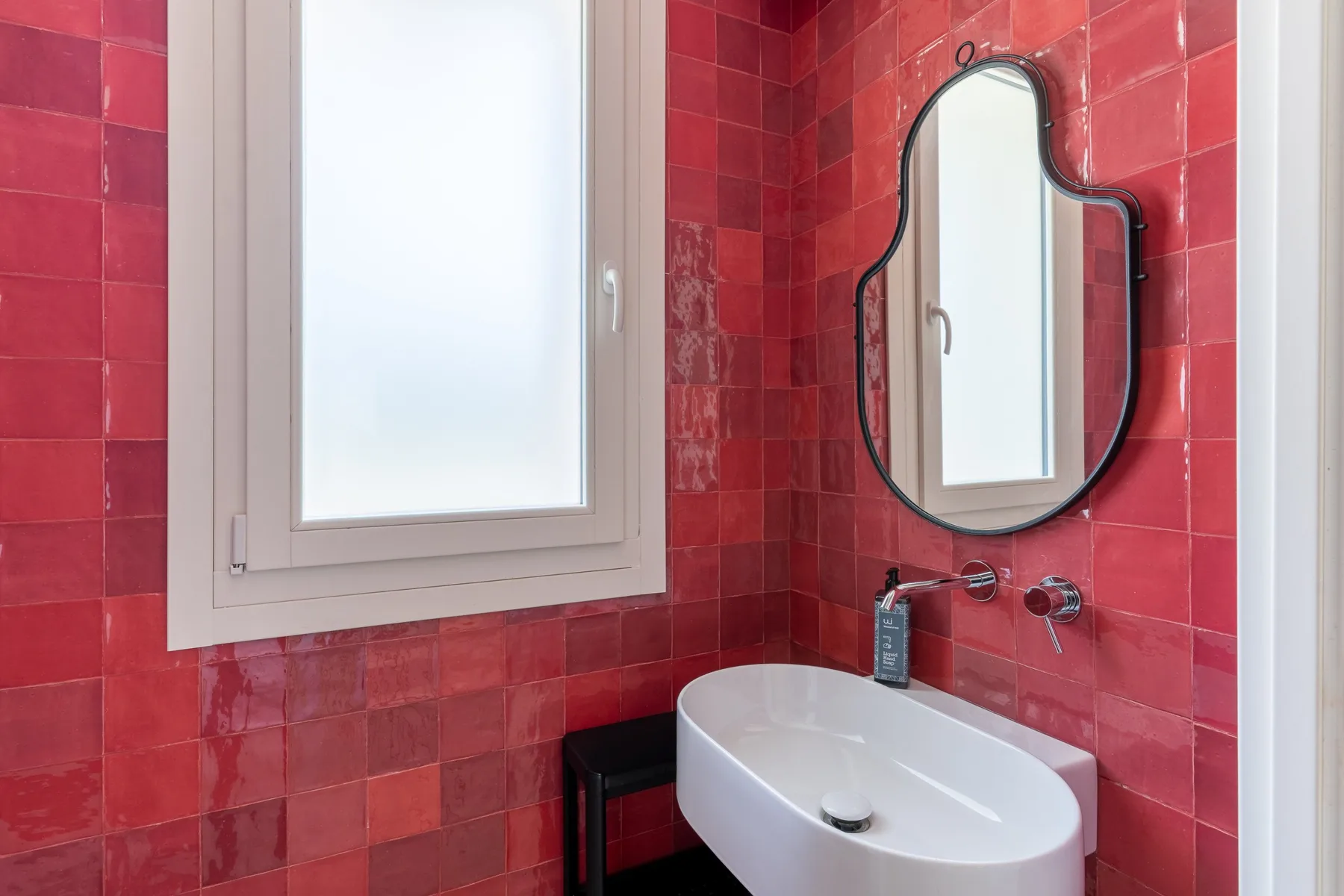 Un baño con lavabo blanco, paredes de azulejos rojos y una ventana.