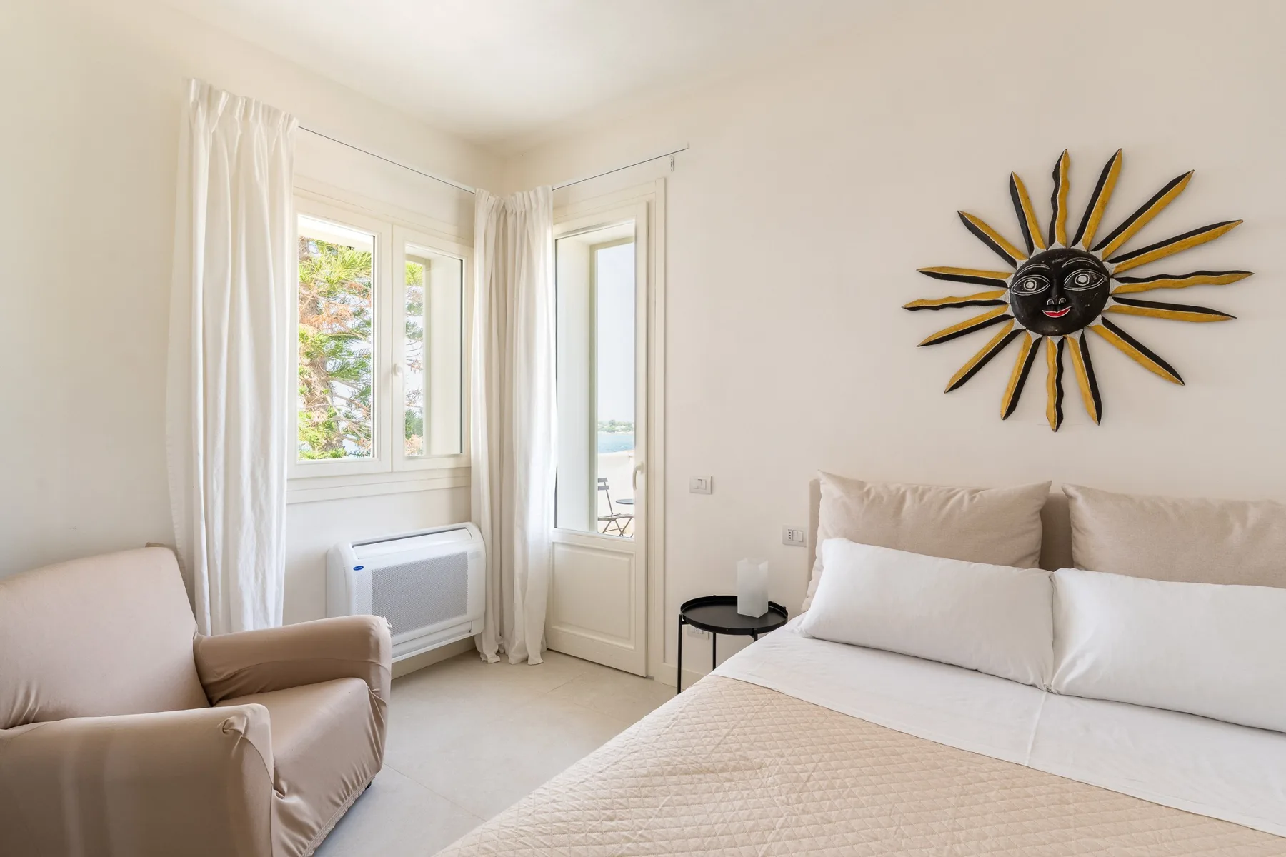 Un dormitorio luminoso con una cama, un sillón y ventanas con cortinas blancas.