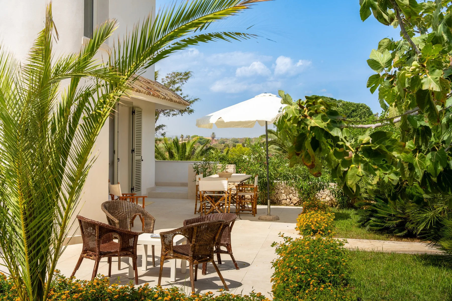 Un patio exterior soleado con una mesa de comedor, una sombrilla y vegetación alrededor.