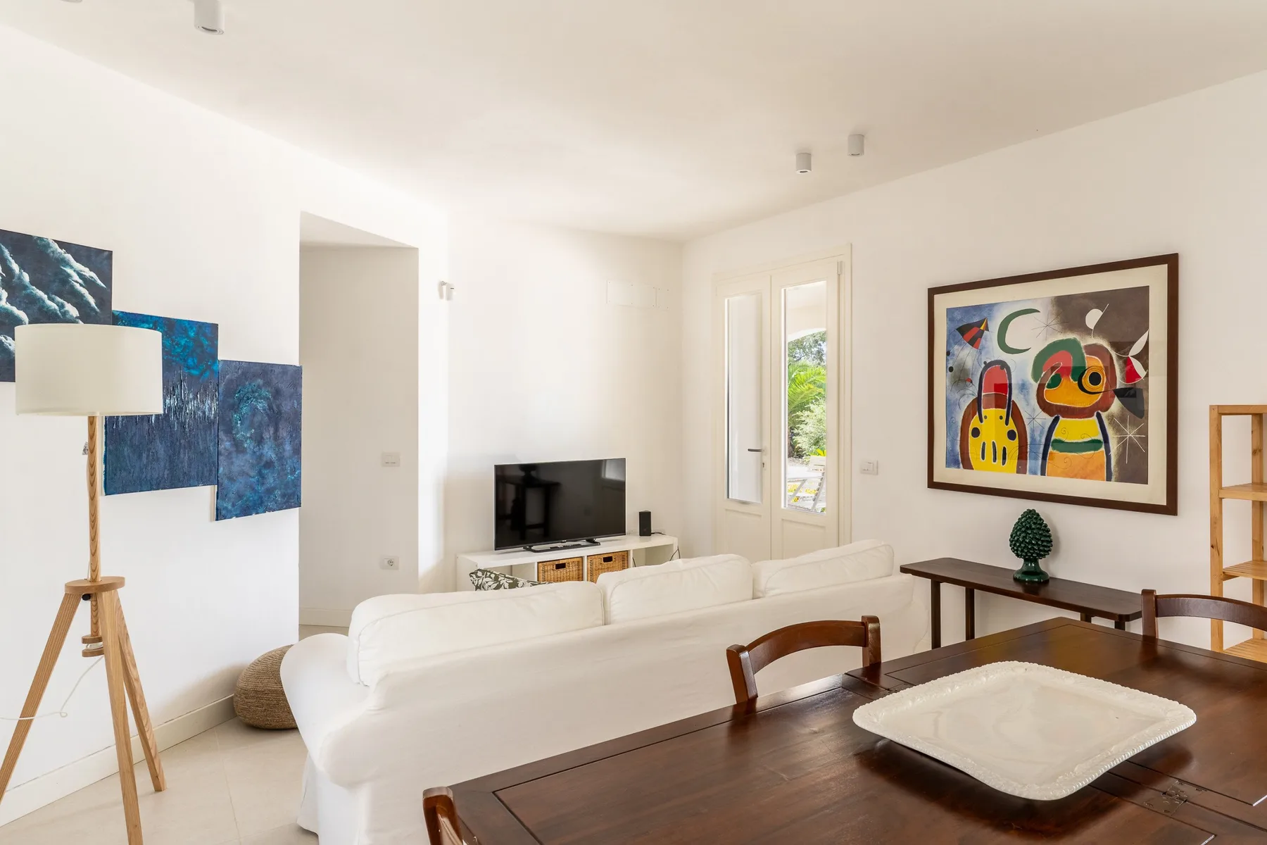 Una luminosa sala de estar con asientos blancos, un televisor y arte colorido en la pared.