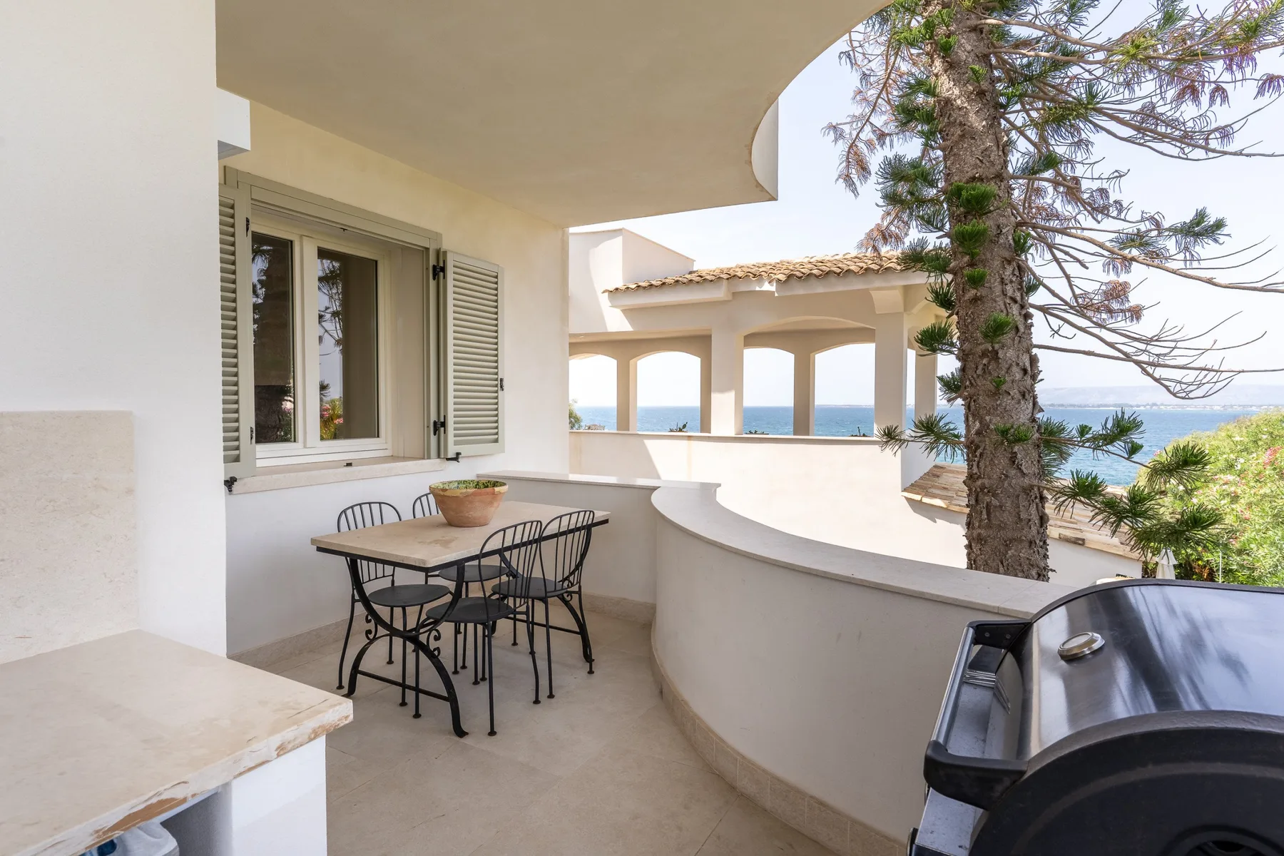 Una terraza cubierta tiene una mesa de comedor junto a un muro curvo, con vistas al mar.