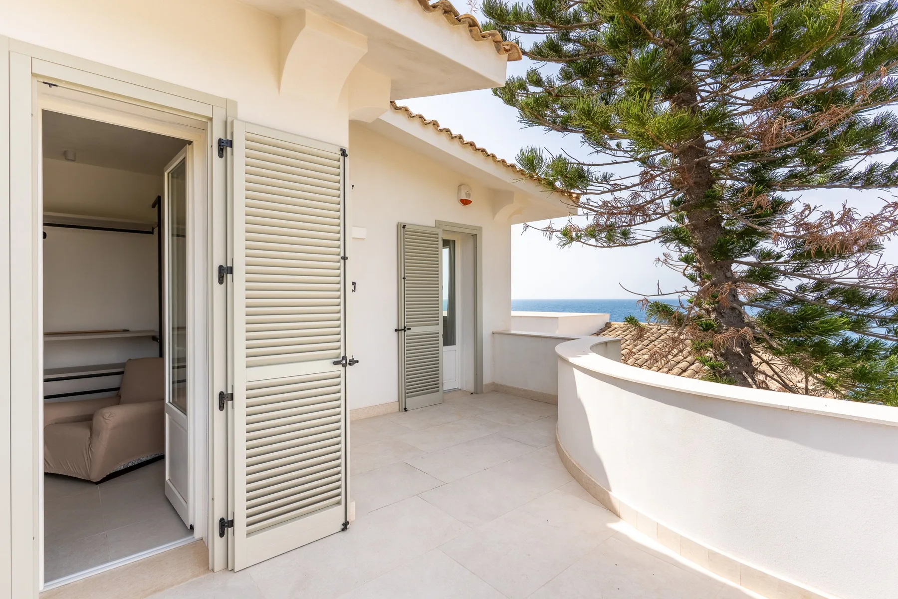 Una terraza exterior con puertas de persiana abiertas, un árbol y vistas al mar.