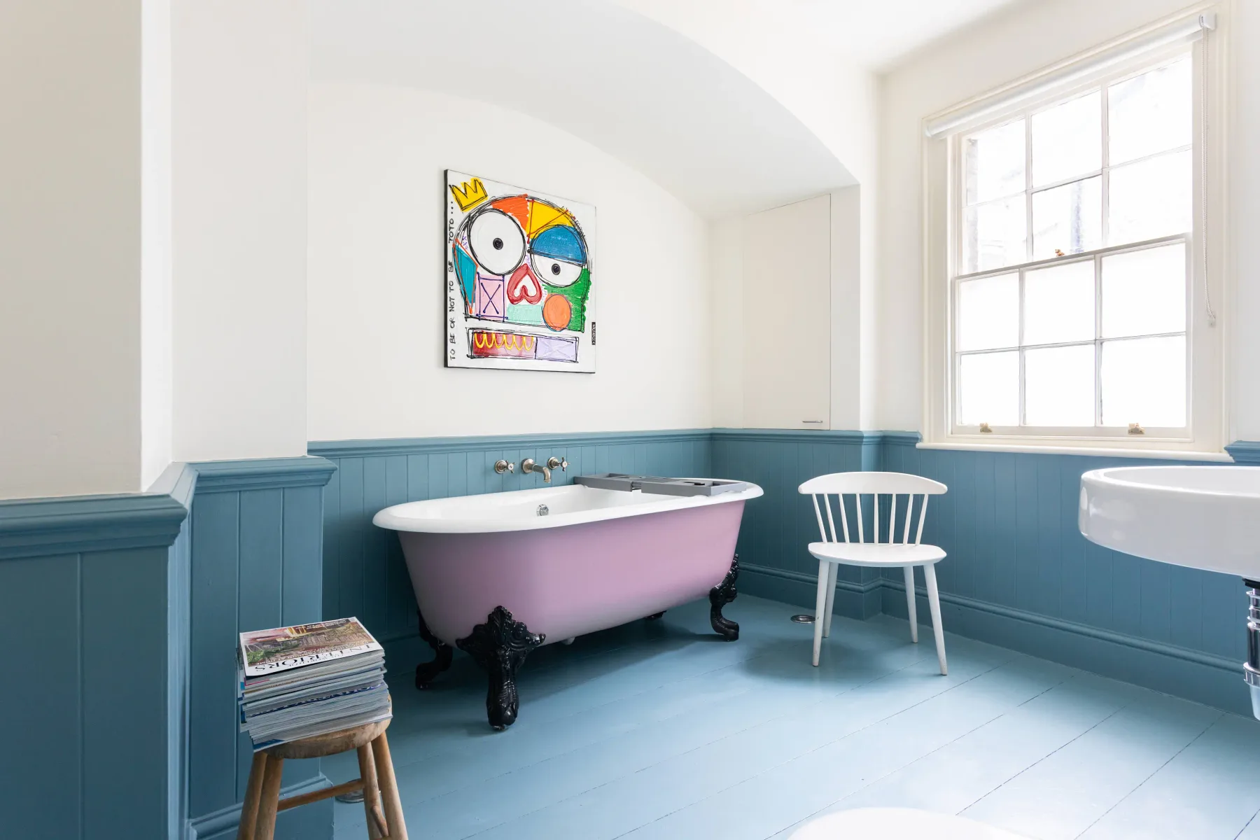 A bright bathroom has a freestanding tub, a chair, and colorful wall art.