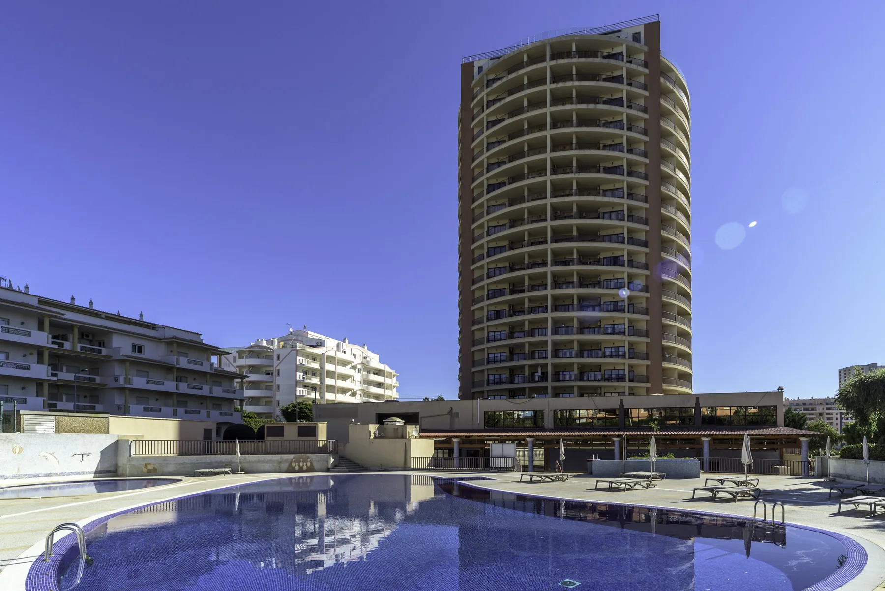 An outdoor swimming pool sits beside several apartment buildings under a clear sky.