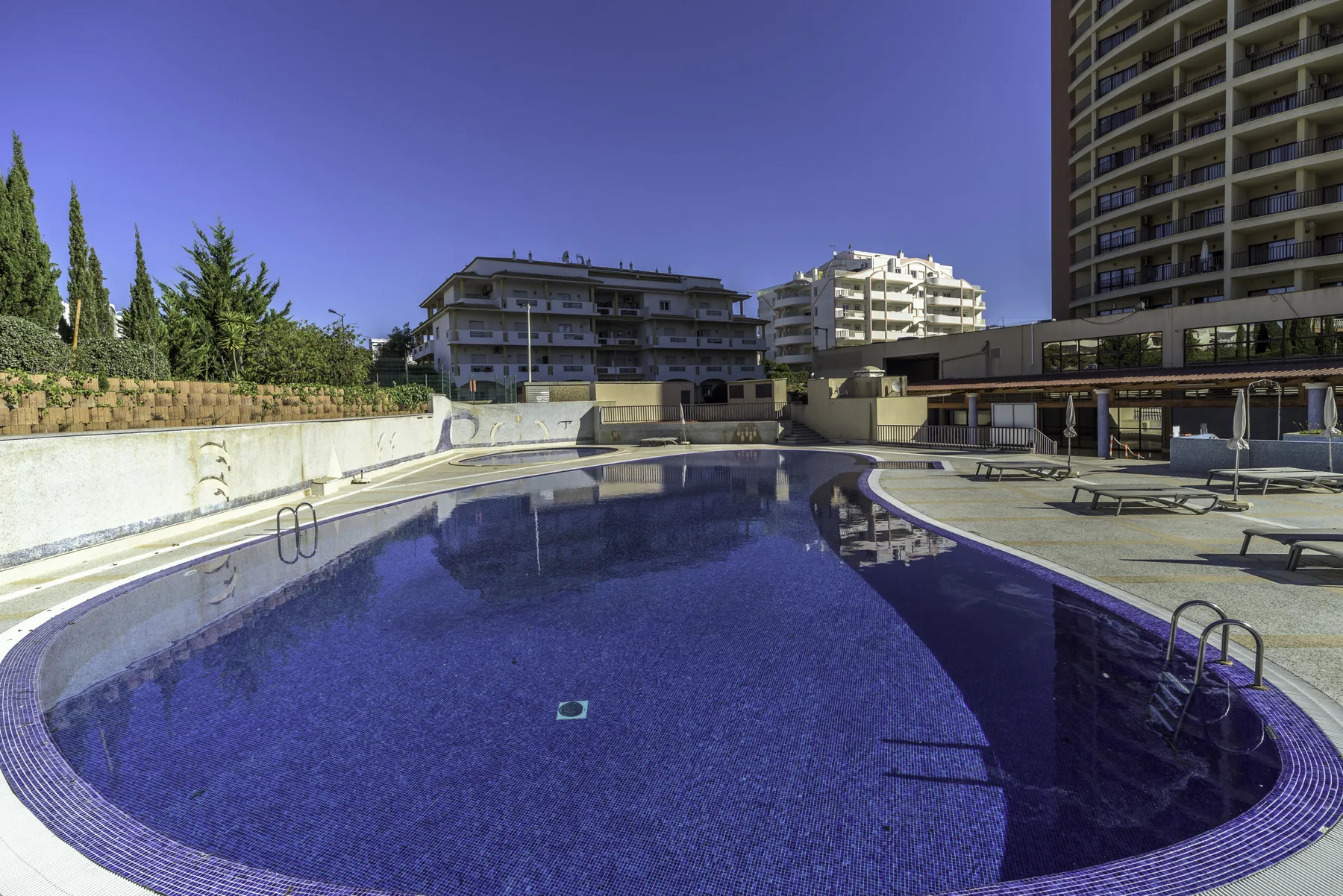 An outdoor swimming pool sits beside a paved deck and surrounding apartment buildings.