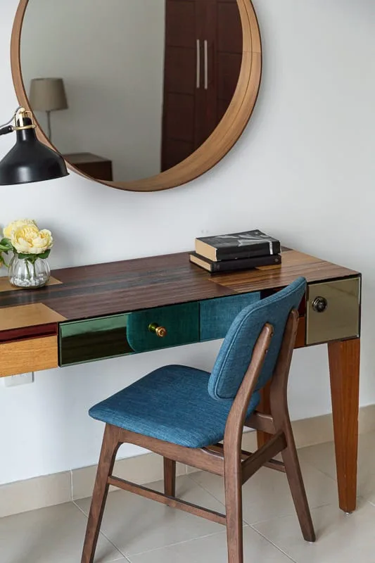 A wooden desk with a blue upholstered chair sits beneath a round mirror.