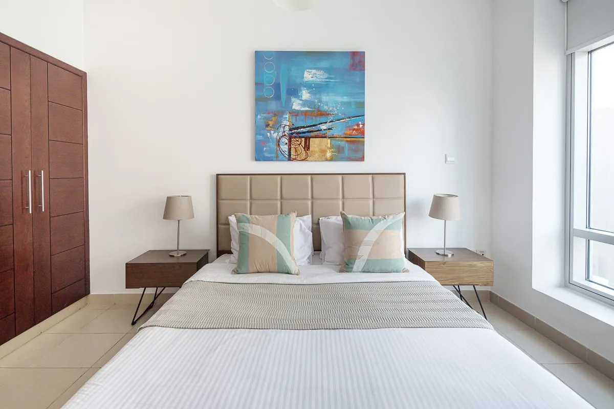 A bright bedroom with a neatly made bed, bedside tables, and colorful wall art.
