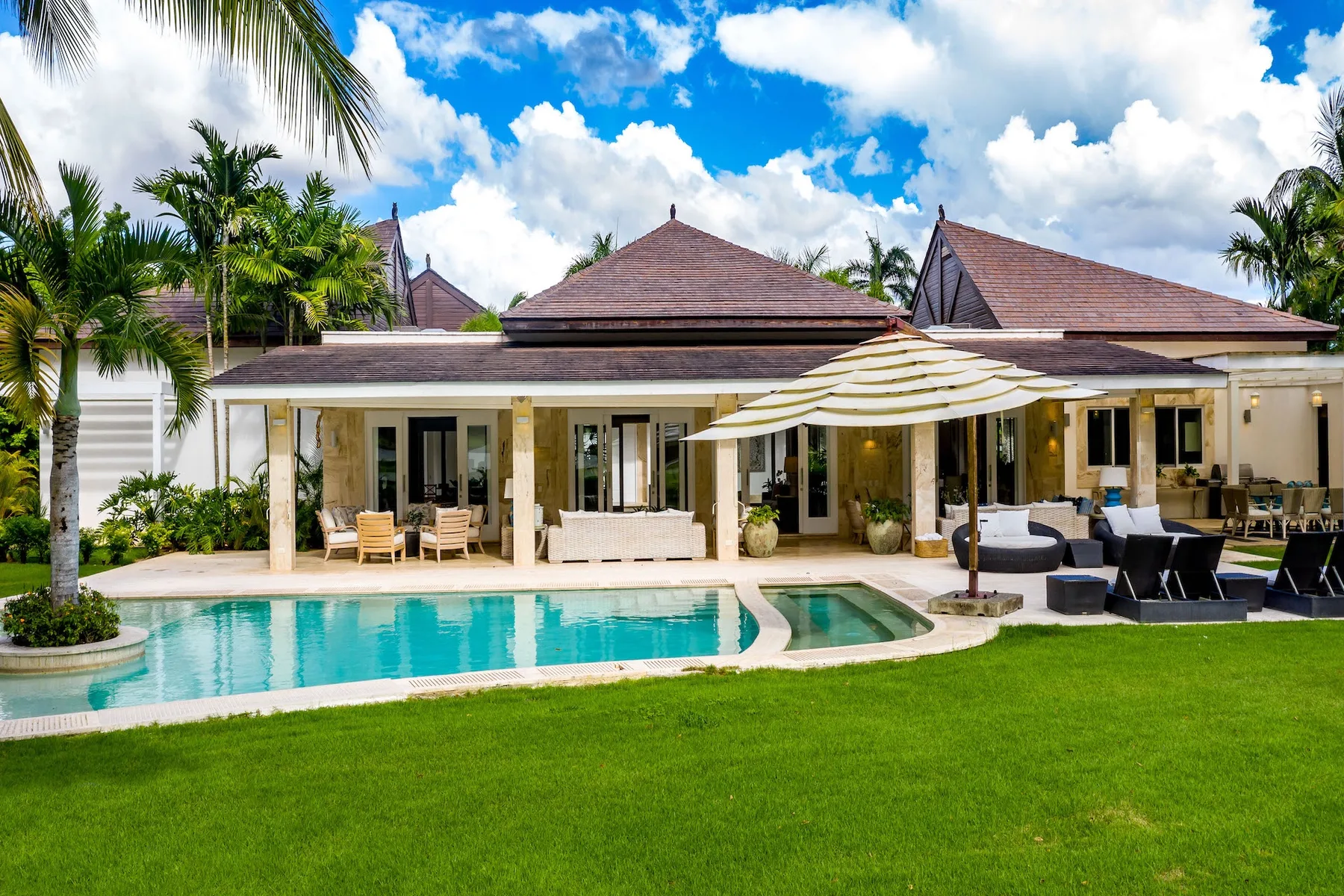 A tropical villa opens onto a swimming pool, with patio seating and palm trees.