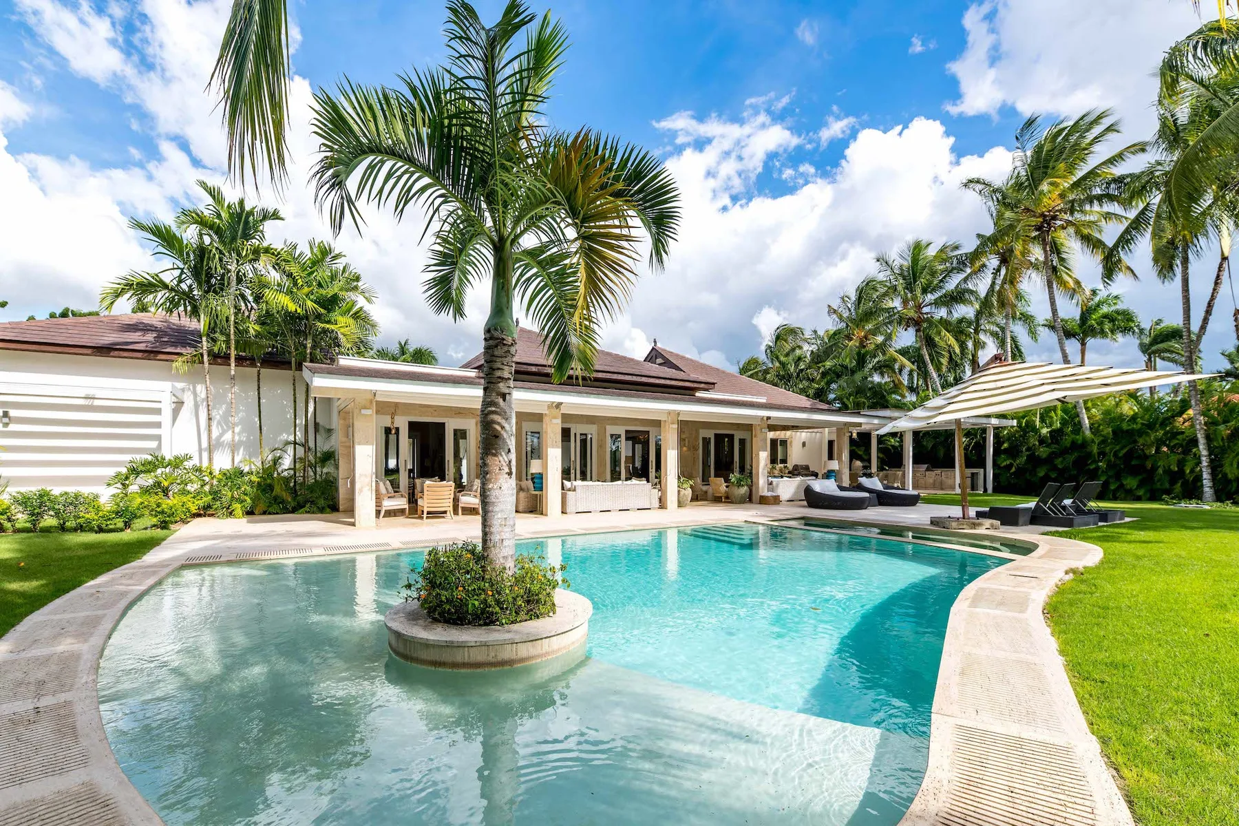 A tropical outdoor pool beside a low-rise building, surrounded by palms and green lawn.