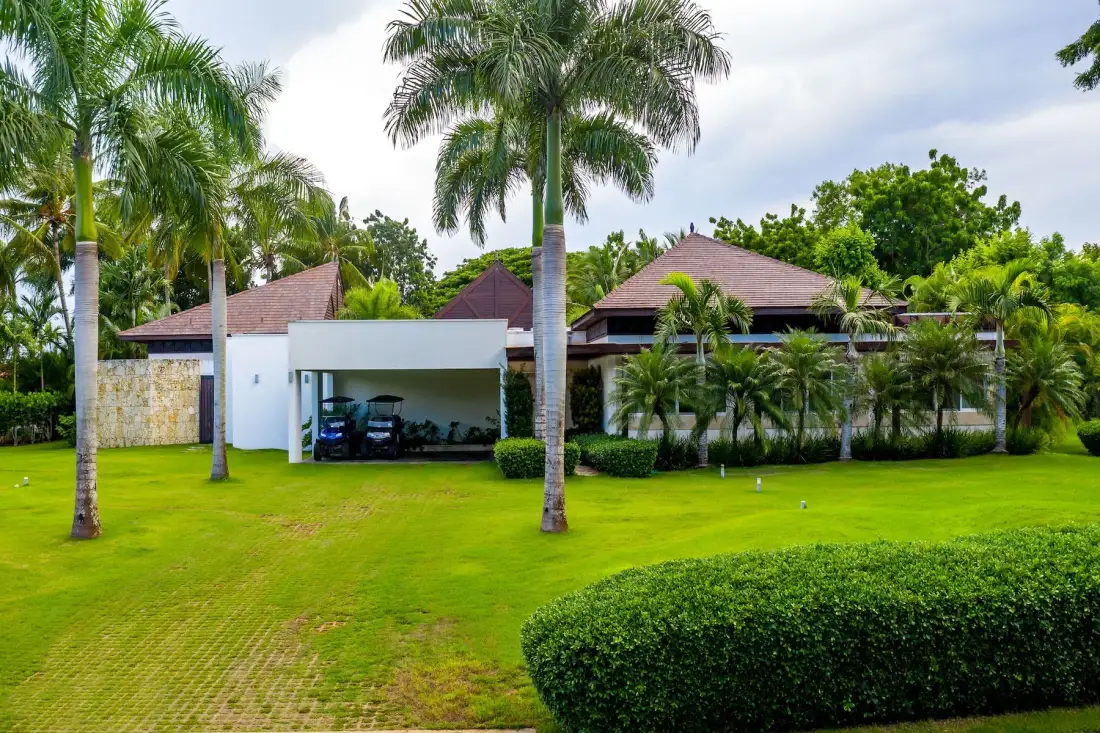 A landscaped property exterior with palm trees, green lawn, and a covered parking area.