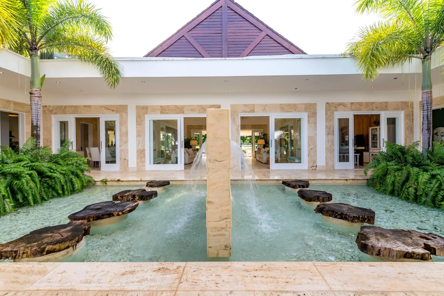 Un patio de estilo resort tiene una fuente poco profunda, plataformas de piedra y vegetación alrededor.