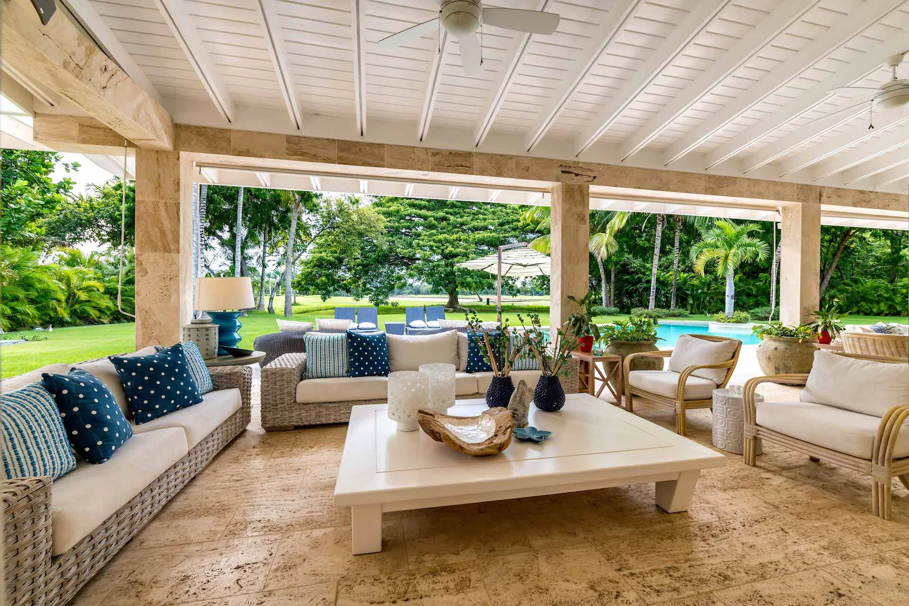 A covered outdoor lounge with cushioned seating and a low table overlooks a garden.