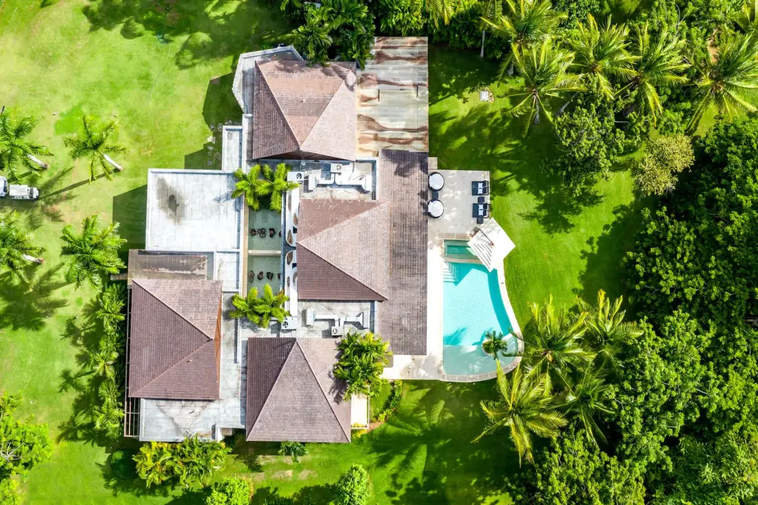 An aerial view shows a house beside a swimming pool, surrounded by greenery.