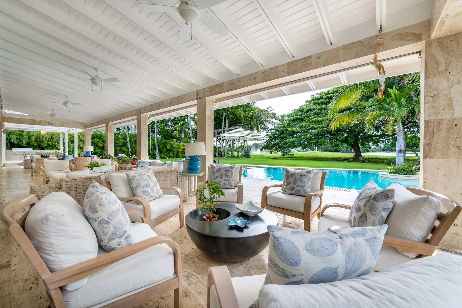 Un salón exterior cubierto con asientos acolchados da a una piscina y un jardín frondoso.