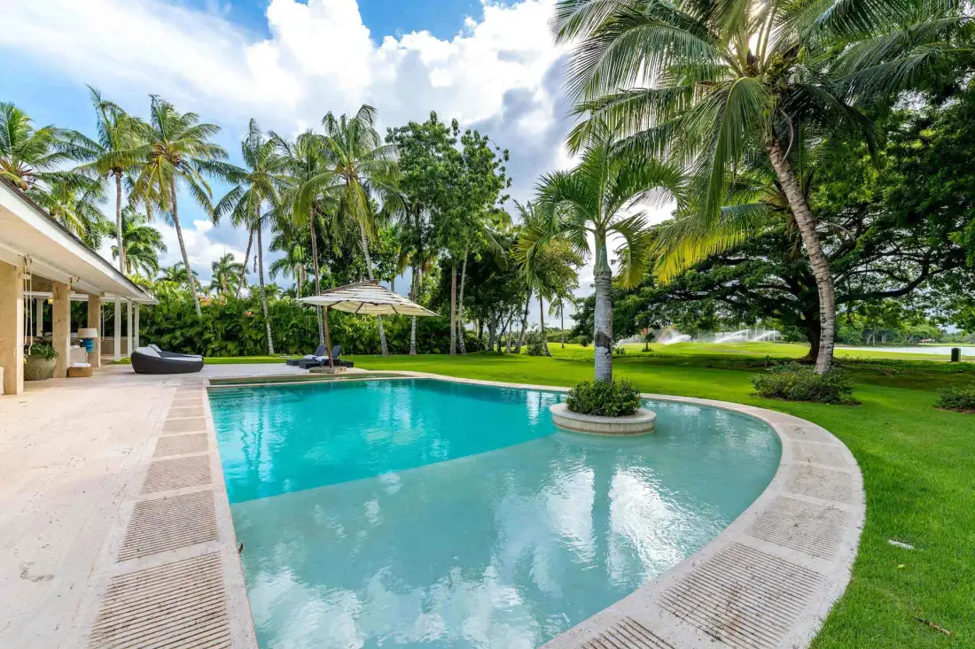 A curved swimming pool sits beside a lawn and palm trees.