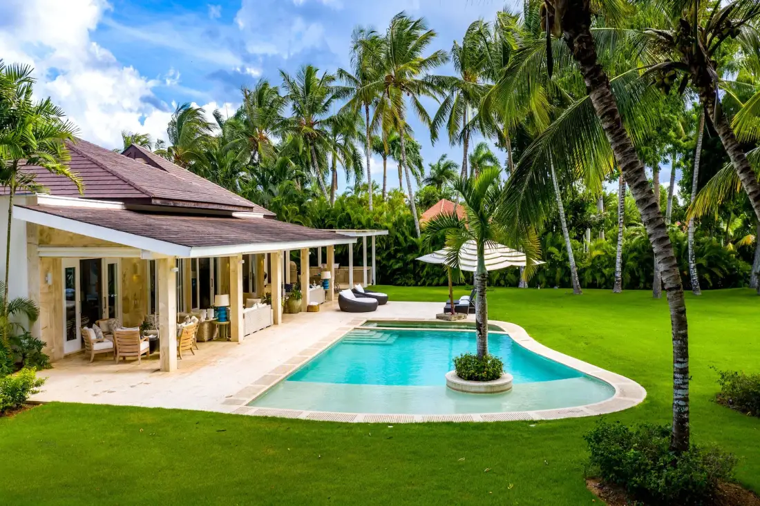 A tropical villa opens onto a swimming pool surrounded by palm trees and lawn.