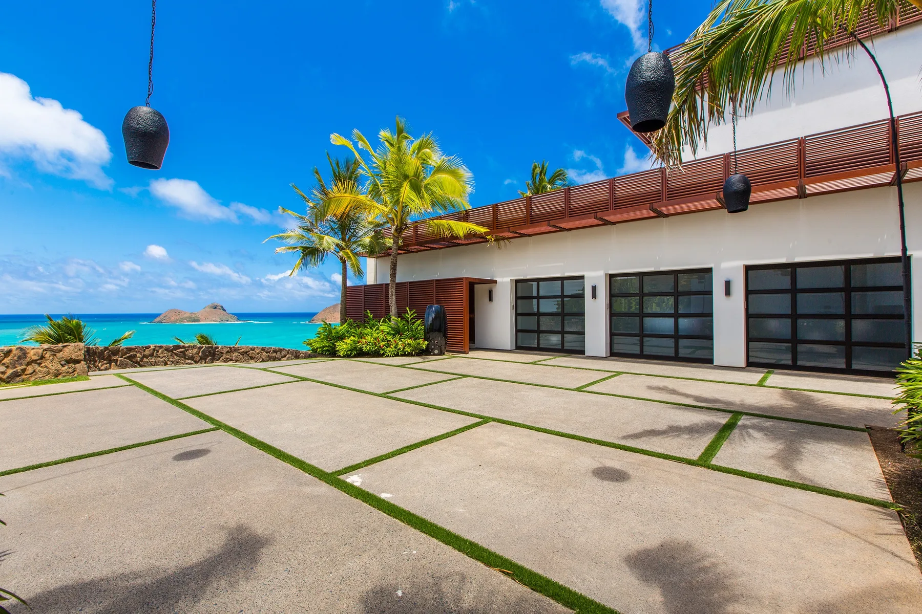 A paved courtyard faces a white building, with palm trees and the ocean beyond.