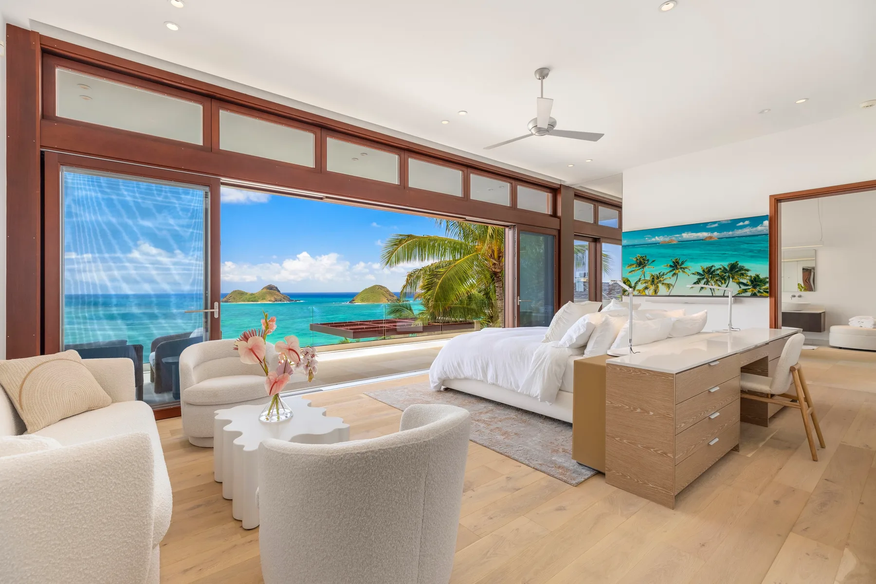 A bright bedroom seating area faces large windows overlooking a tropical beach.