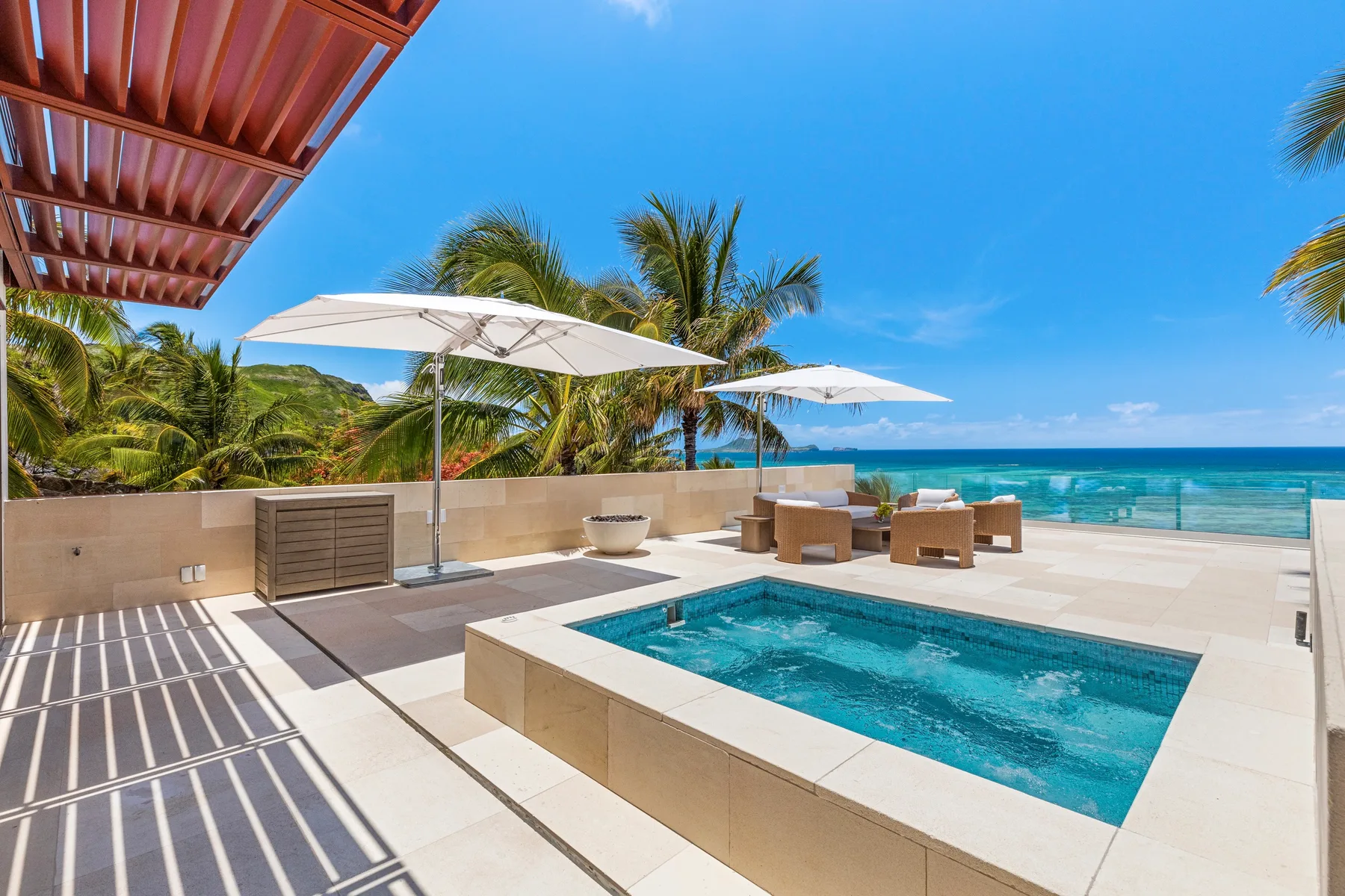 A sunlit pool terrace overlooks the ocean, with umbrellas and palm trees nearby.
