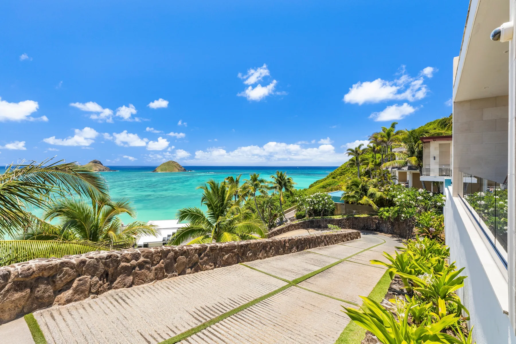 A paved seaside terrace overlooks turquoise water, with palm trees and a rocky shore.