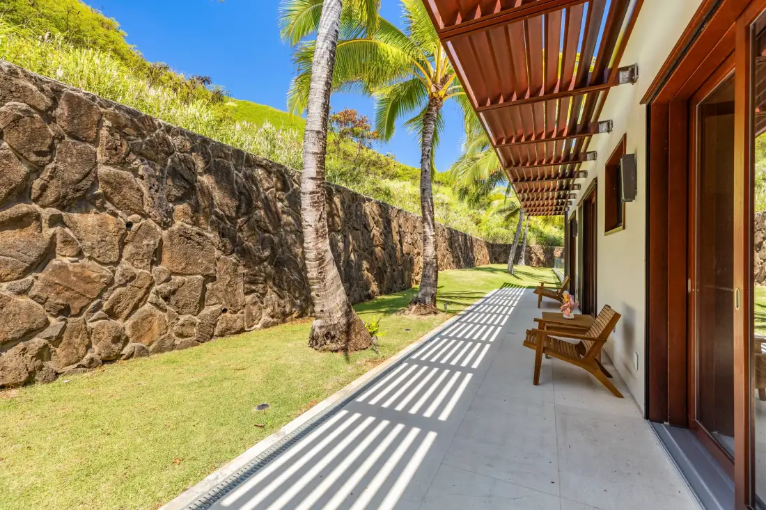 A covered patio with wooden seating overlooks a grassy area and stone wall.
