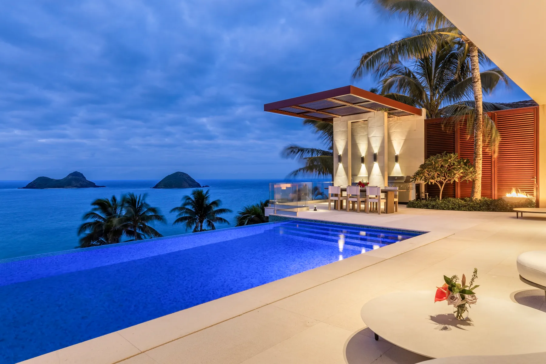 An illuminated swimming pool and patio overlook the ocean at dusk.