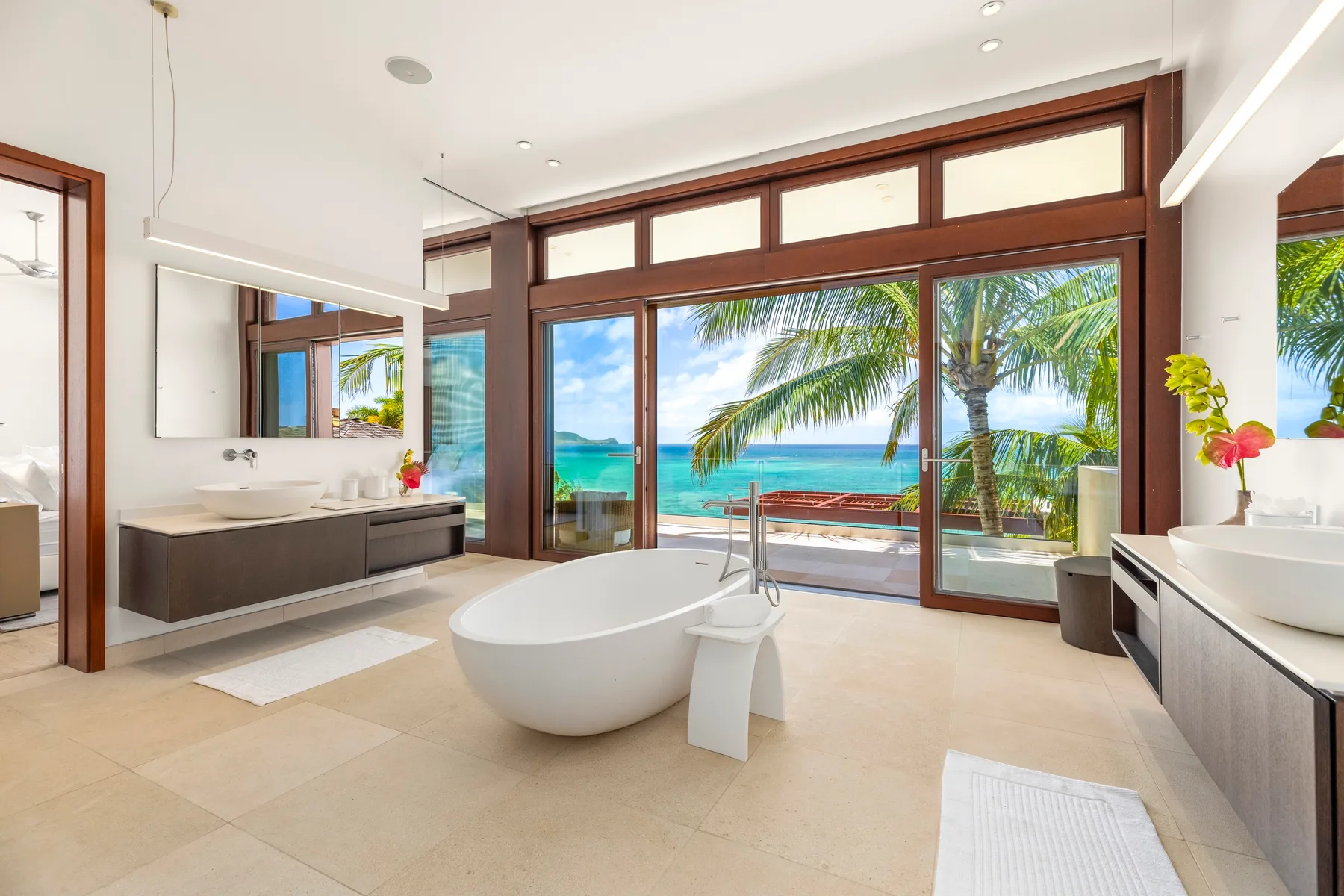 A bright bathroom with a freestanding tub and large windows overlooking palm trees and the sea.