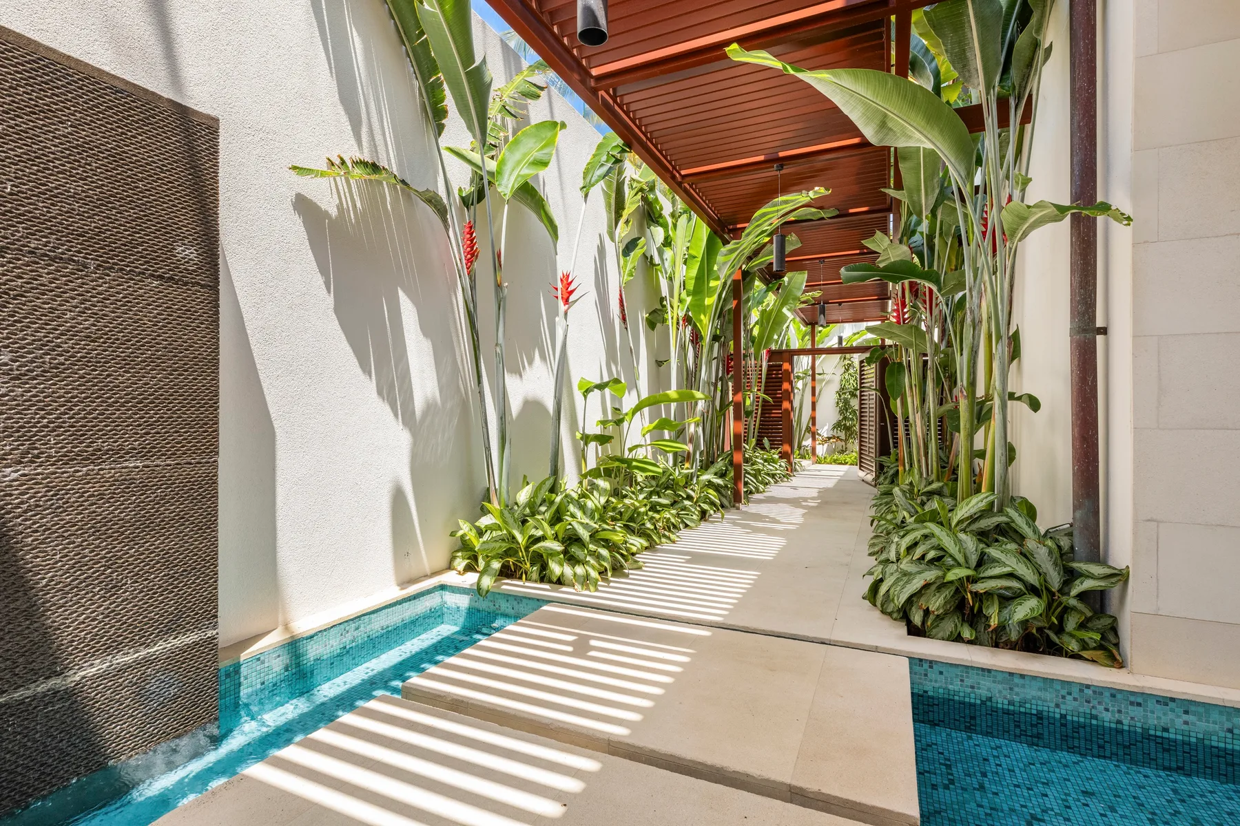 Un pasillo exterior soleado discurre entre piscinas estrechas y exuberantes plantas tropicales.