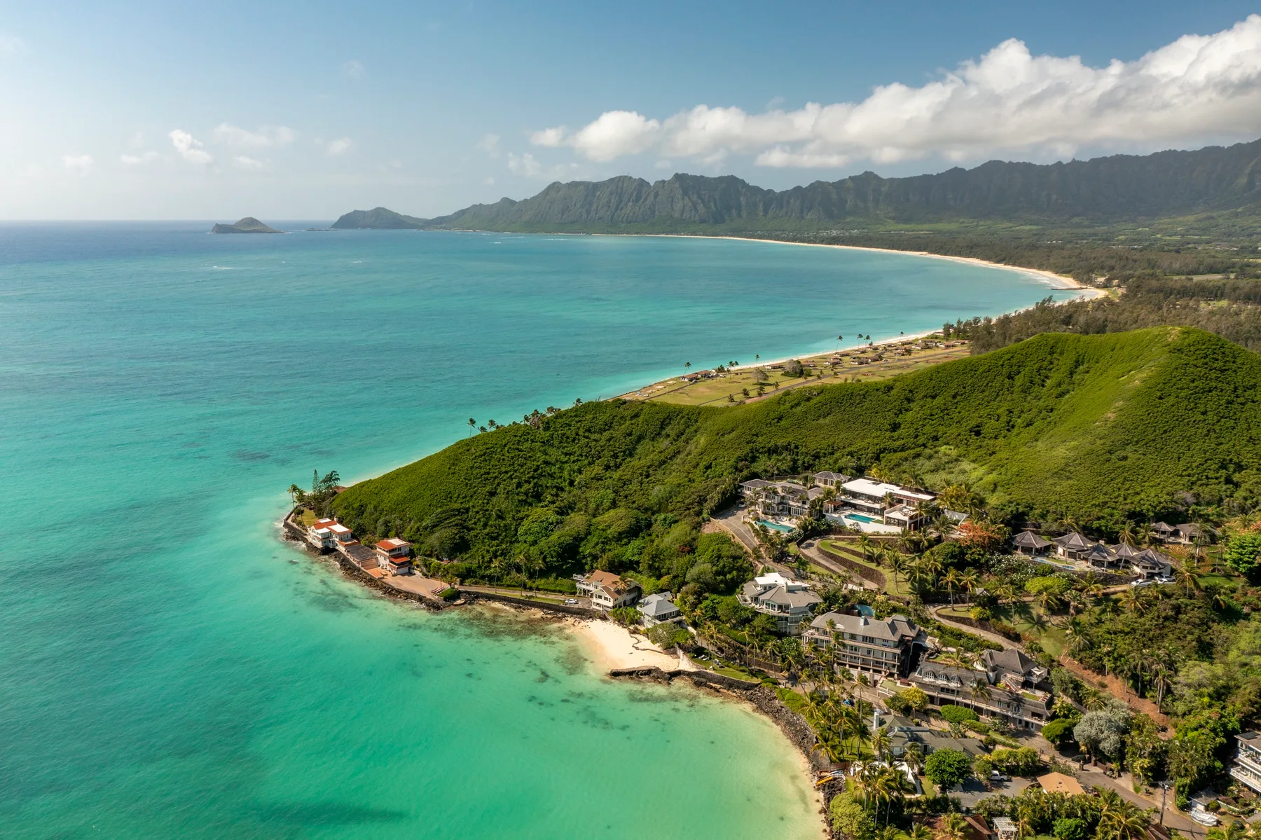 Aerial view of a tropical coastline with turquoise water, green hills, and scattered buildings.