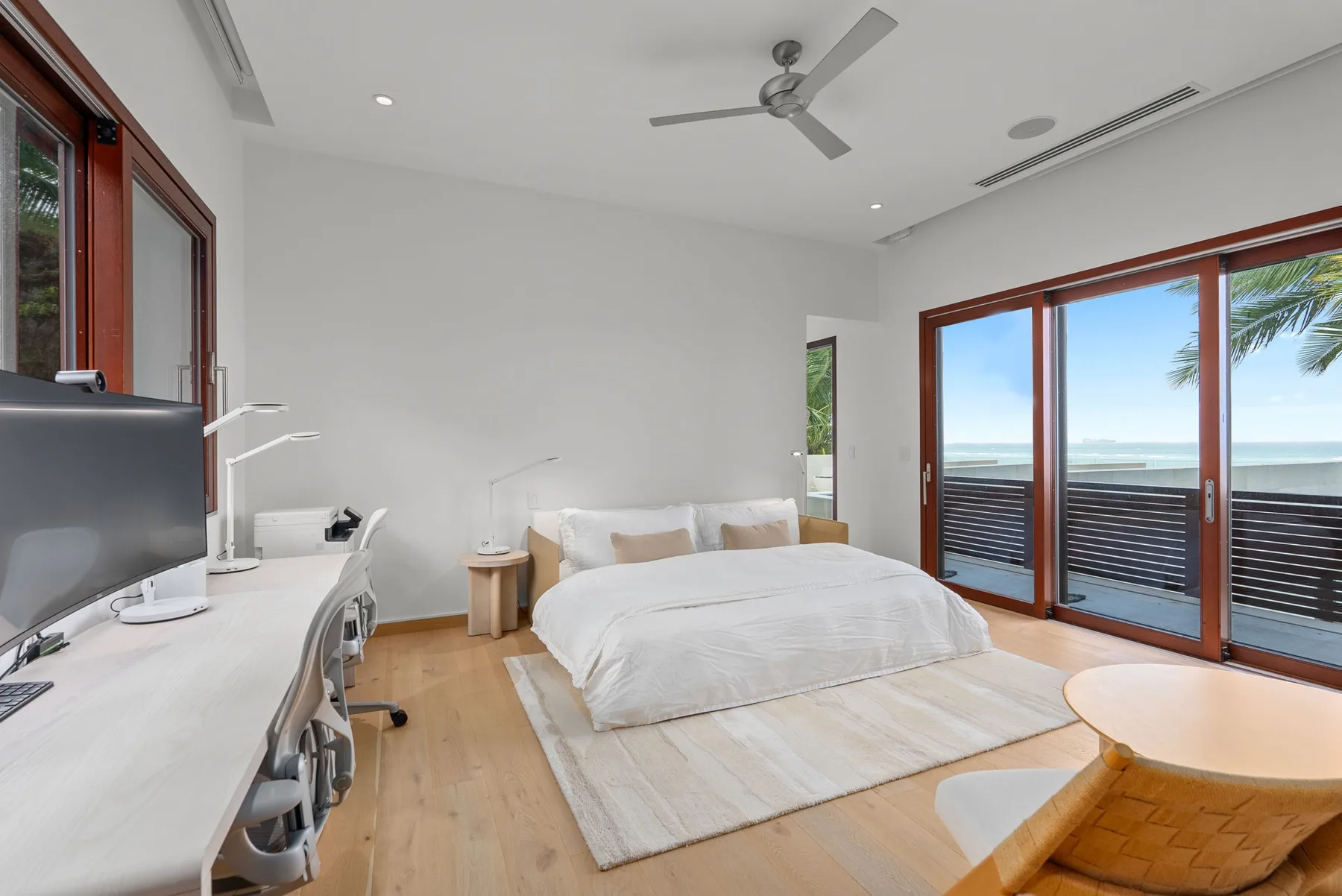 A bright bedroom with a bed, desk, and large windows overlooking the ocean.