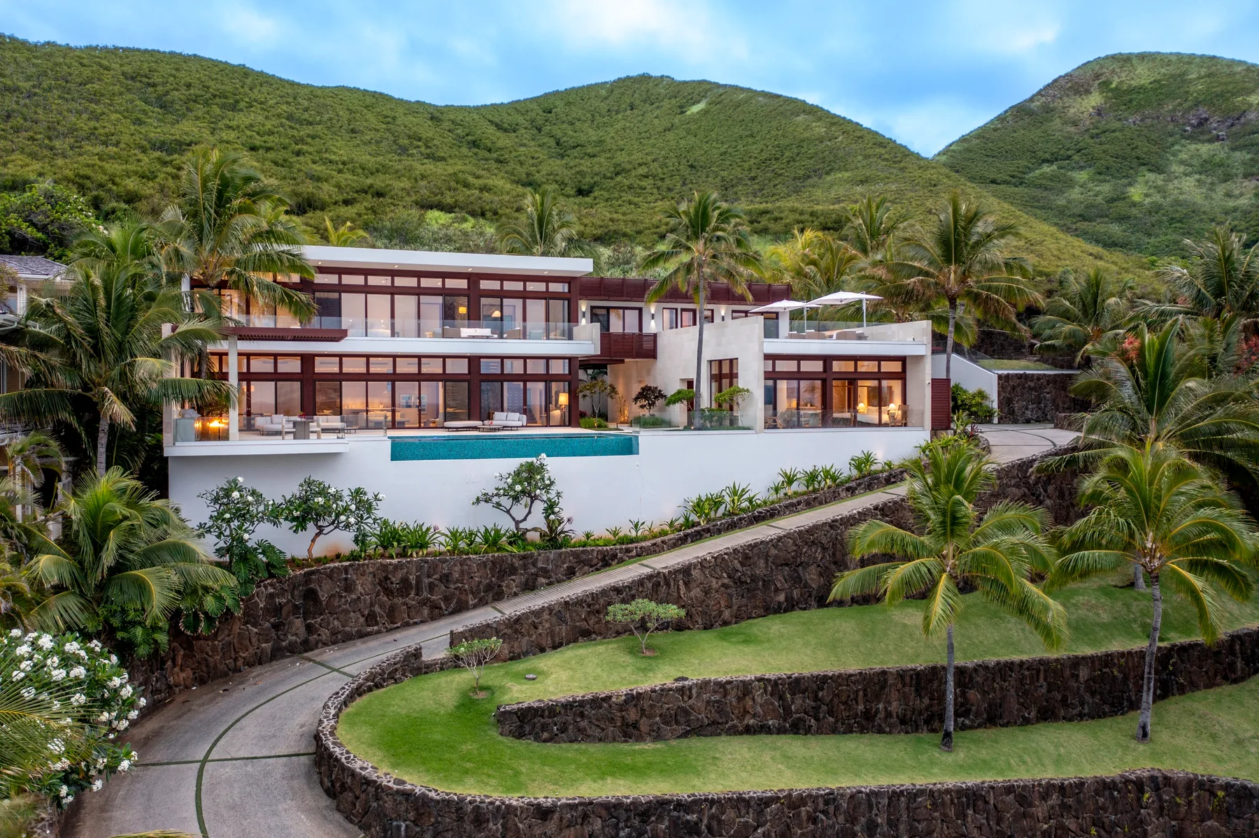 A modern hillside house with a swimming pool, palm trees, and green mountains.