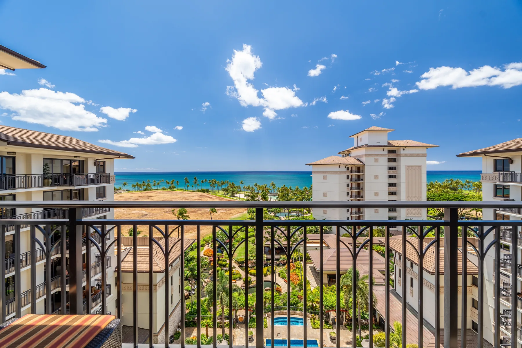 A balcony overlooks nearby buildings, a pool, and the ocean beneath a blue sky.