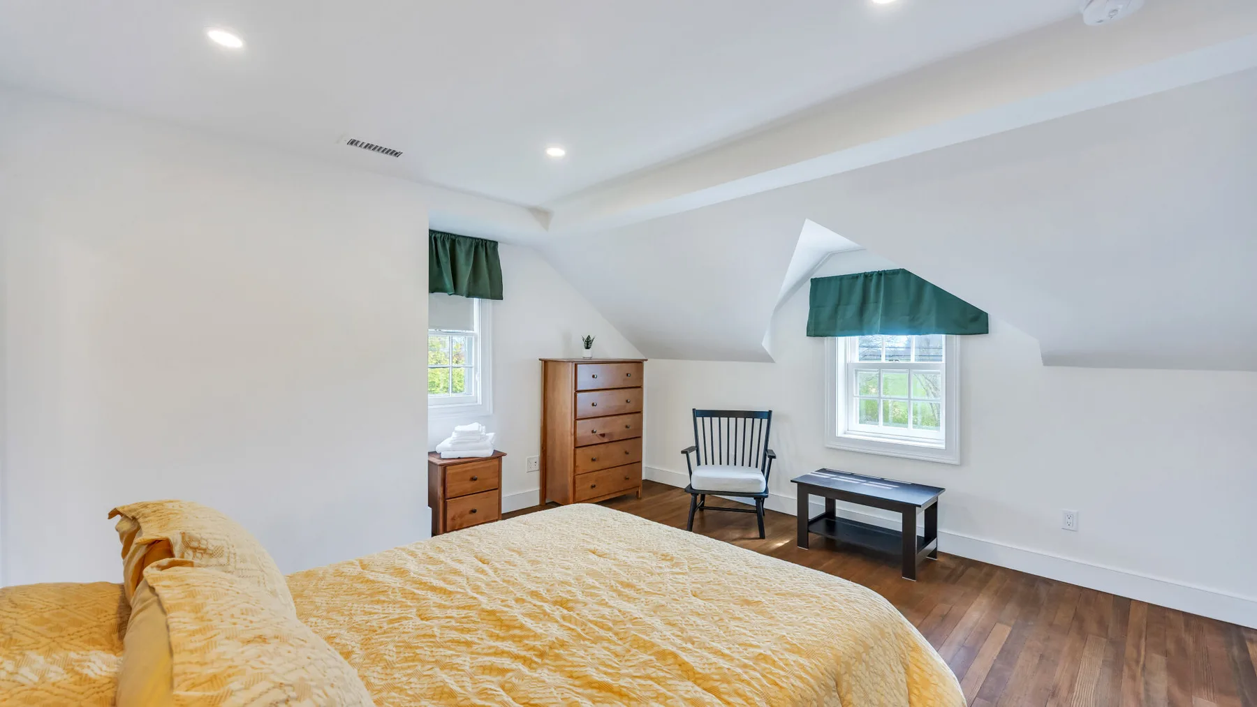 A bright attic bedroom with a bed, dresser, and small seating area.