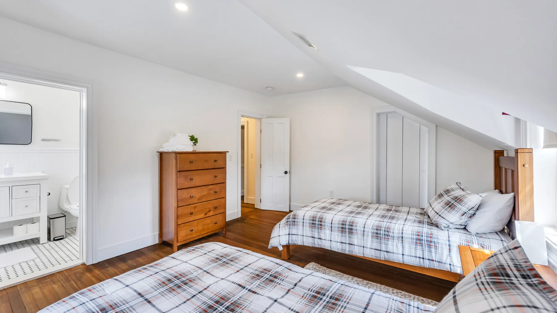 A bright attic bedroom with beds, a wooden dresser, and an open doorway.