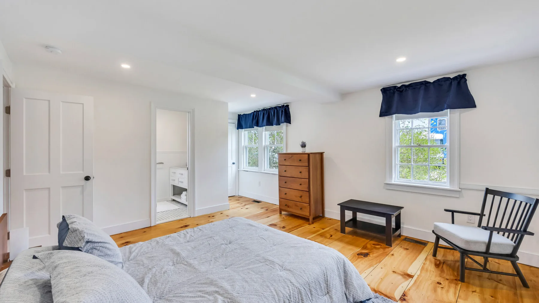 A bright bedroom with a bed, wooden dresser, and windows.