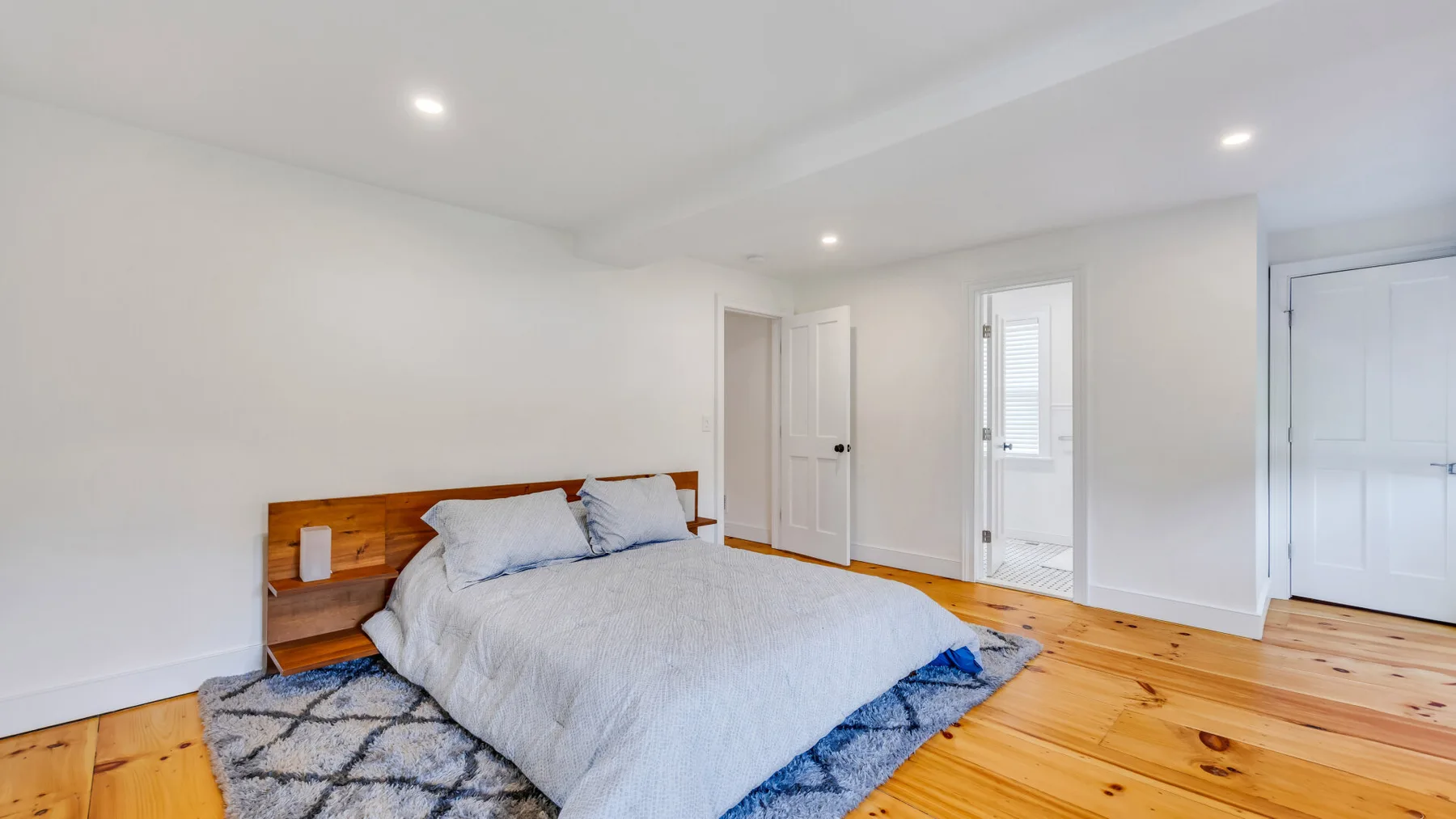 A bright bedroom with a bed on a patterned rug and wooden flooring.