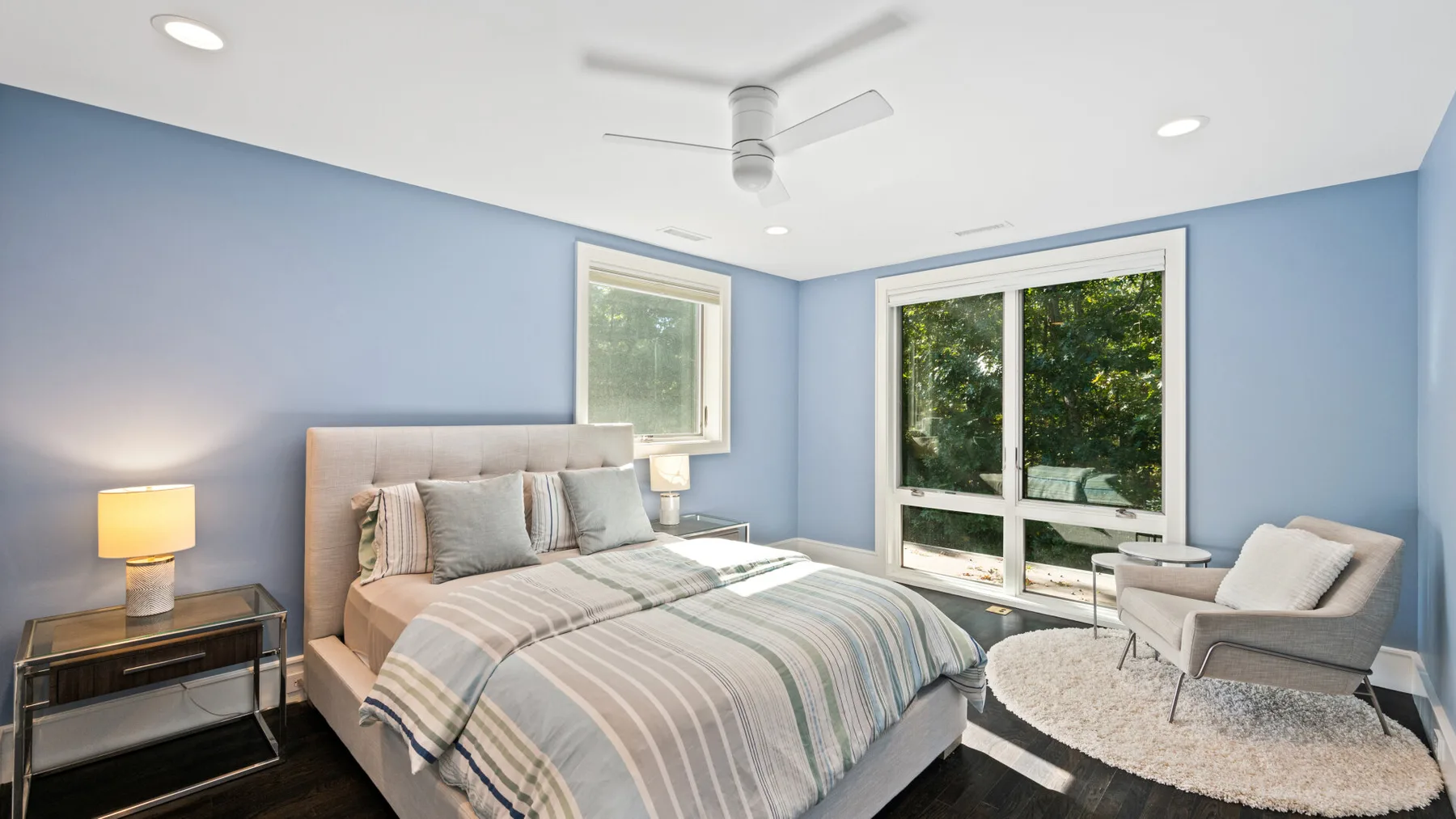 A bedroom with a neatly made bed, blue walls, and large windows overlooking greenery.