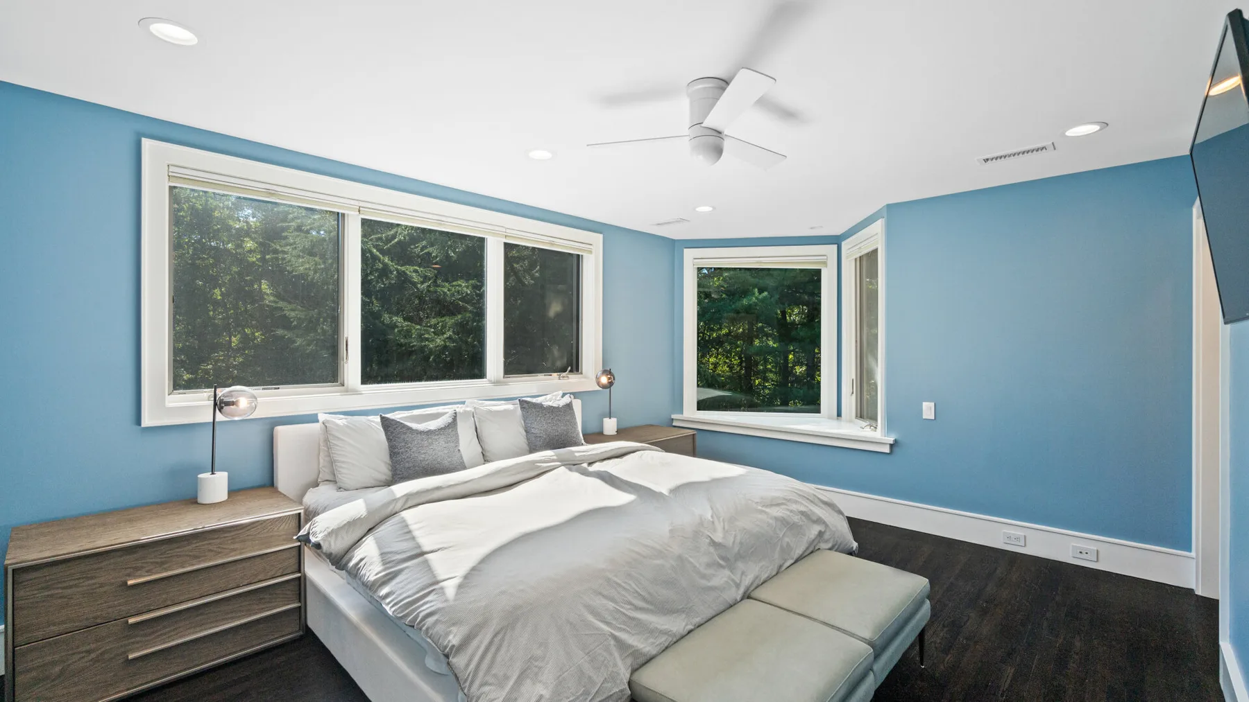 A bedroom with a bed, blue walls, and large windows overlooking greenery.